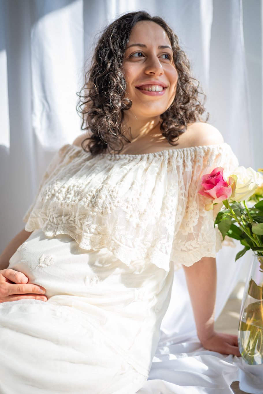 Schwangere Frau in einem weißen Kleid, lächelt mit Blumen im Hintergrund.