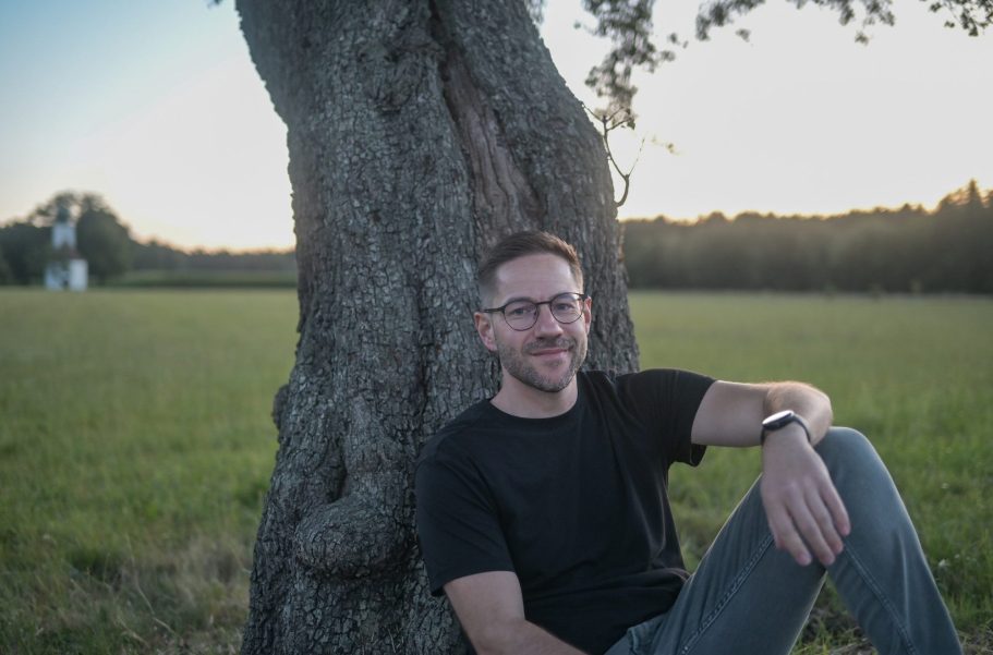 Tobias Luber sitzt lächelnd an einem Baum in einer grünen Landschaft.