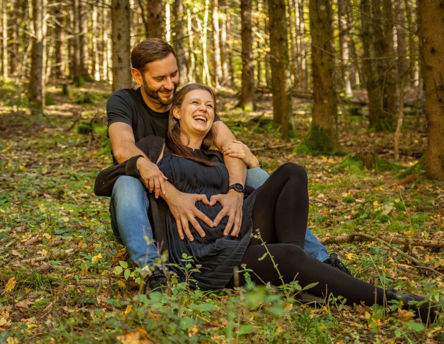 Ein Paar sitzt im Wald, der Mann umarmt die Frau, die schwanger wirkt und lächelt.